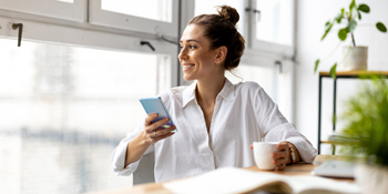 Business woman drinking coffee and smiling as she looks out the window while managing her St. Mary’s Bank account on her phone